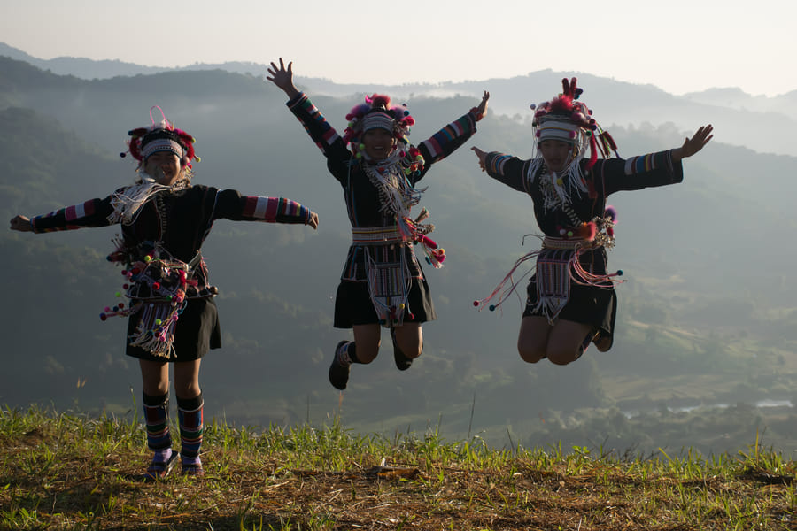 Fotografía de tres personas pertenecientes a la etnia Akha, vistiendo sus trajes tradicionales coloridos y ornamentados, saltando con entusiasmo en un entorno natural montañoso y neblinoso. Transmite alegría, autenticidad cultural y conexión con las tradiciones locales.